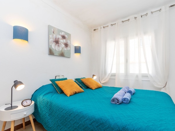 A bedroom with a teal bedspread, two pillows, a bedside lamp, a clock, and a painting on the wall.