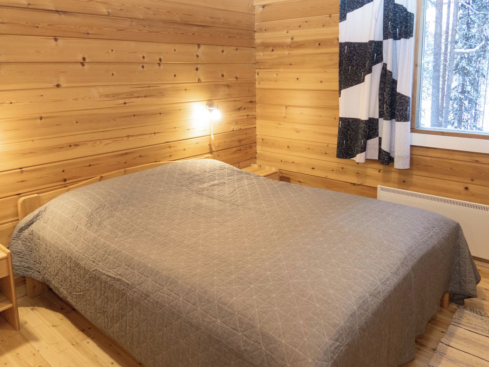 Une pièce de cabane en bois comprenant un lit, une fenêtre donnant sur la forêt et un luminaire fixé au mur.