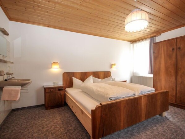 A wooden bed with a headboard, two bedside tables, a nightstand, a wooden wardrobe, a bathroom with a sink and mirror, and a window with curtains.