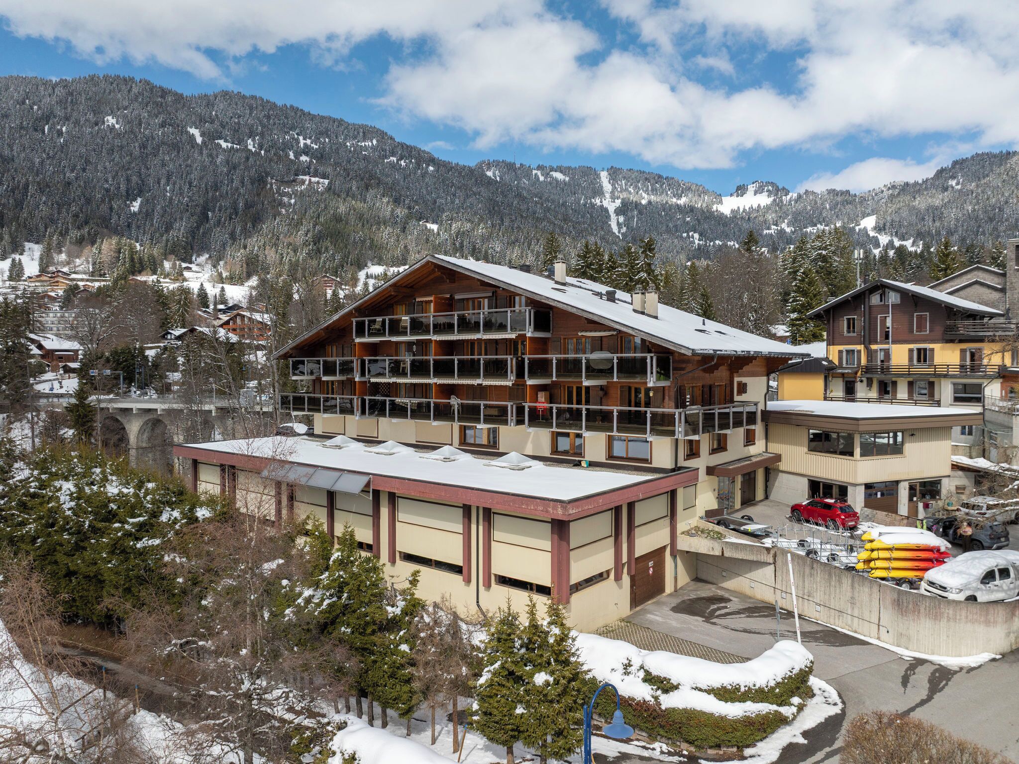Um edifício de vários andares com varandas, uma área de estacionamento com veículos e montanhas cobertas de neve ao fundo.