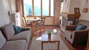 A living room with a sofa, armchair, coffee table, and fireplace. There is a dining table with chairs and a view of mountains through large windows. - La Vire D'Aufalle 14 Inférieur by Interhome (Alpe des Chaux)