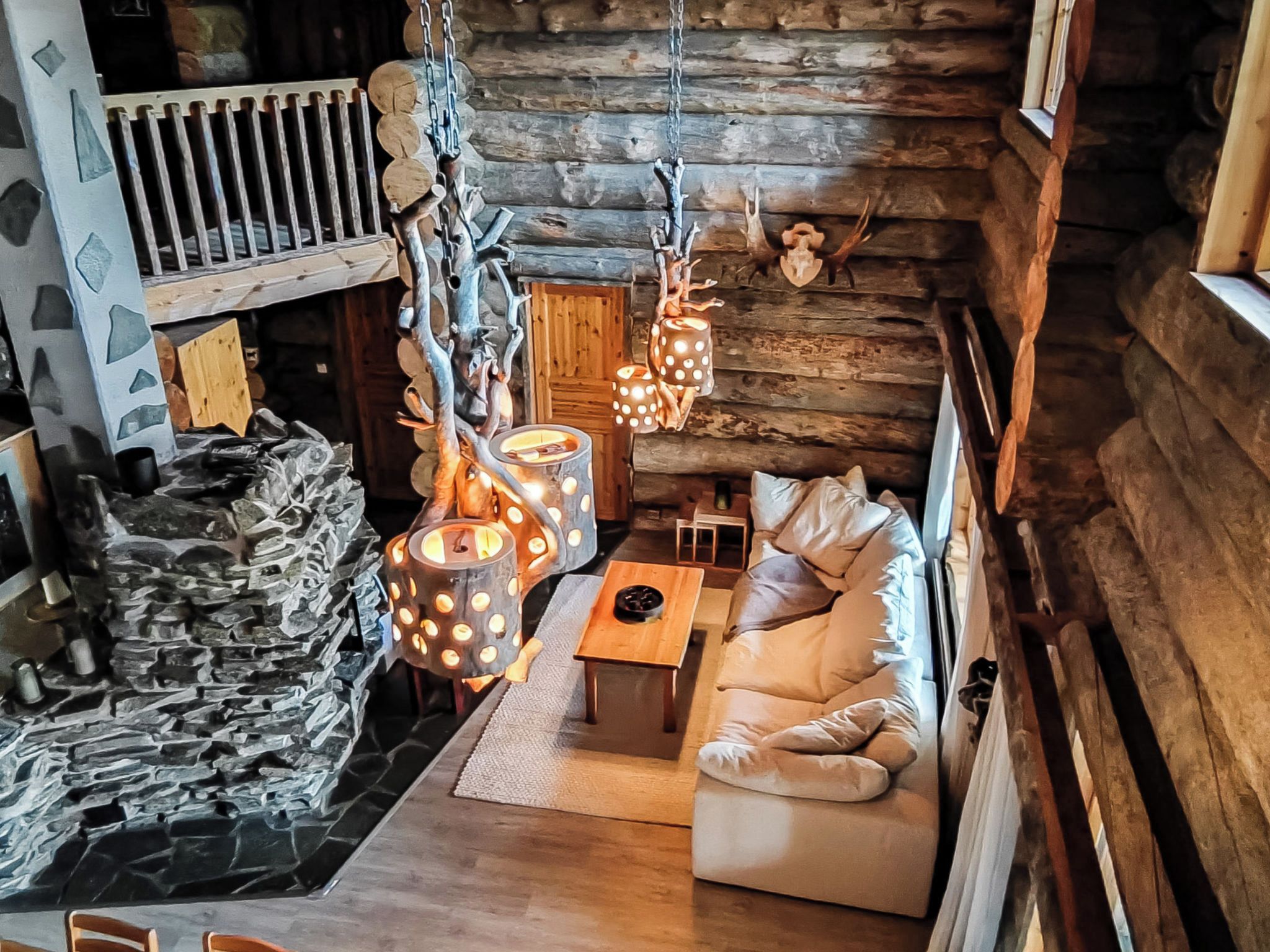 Interior de una cabaña de troncos con pared de piedra, escalera de madera, sofá e iluminación colgante.
