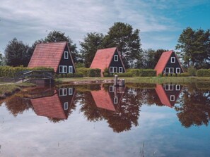 Three traditional houses with red roofs and white window frames, reflected in a calm body of water, surrounded by greenery and trees.