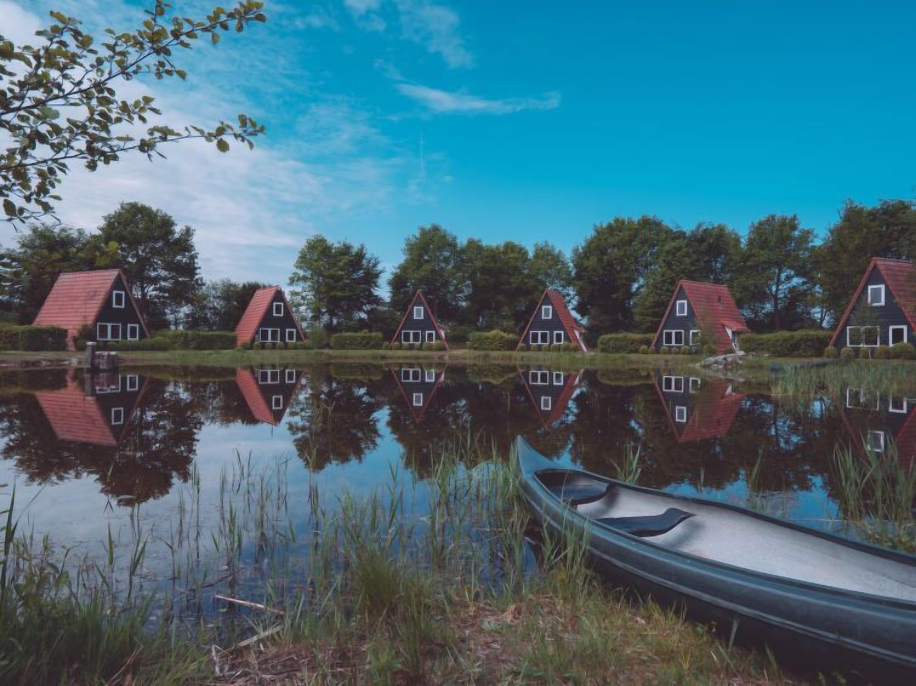 En række traditionelle huse med røde tage ved en rolig sø, en kano i forgrunden og træer i baggrunden.