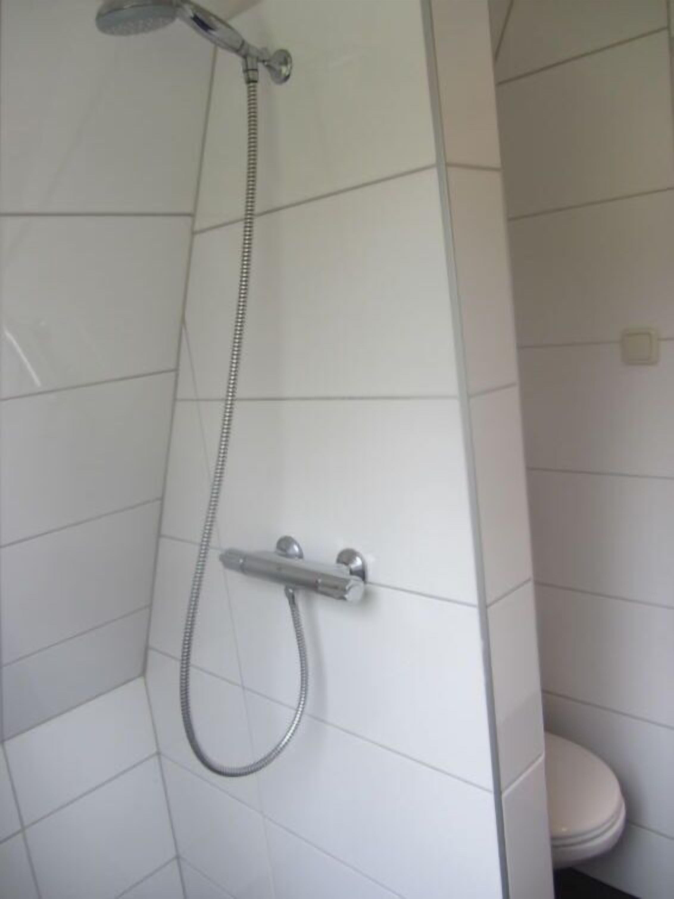 Une salle de bain avec des murs carrelés de carreaux blancs, une douche avec un pommeau de douche et un flexible, et des toilettes.