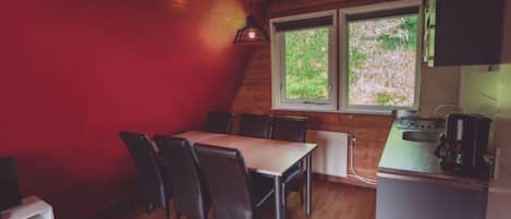 A cozy dining area with a wooden table and chairs, a red accent wall, and a window with a view of greenery.