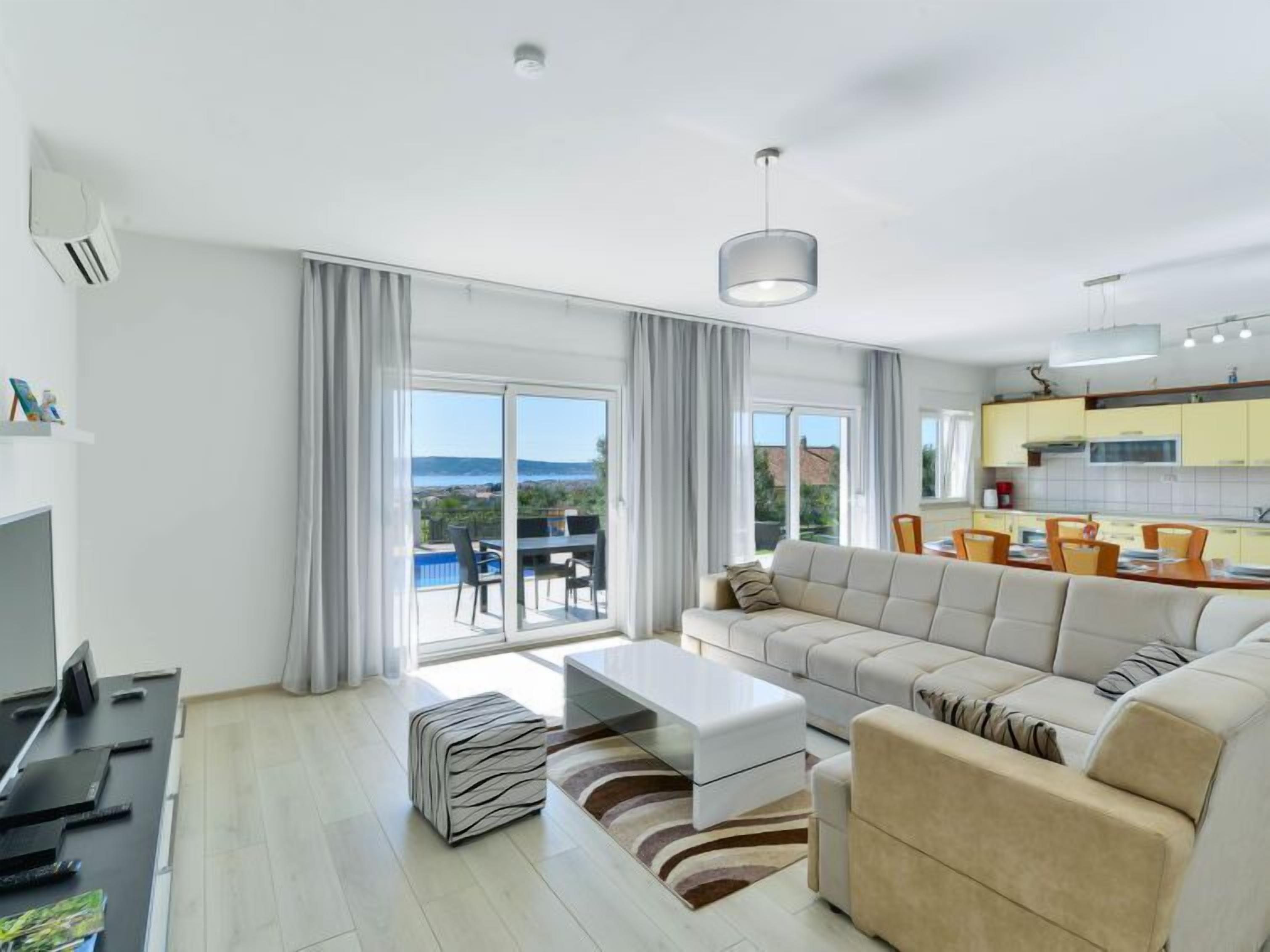 Ein modernes Wohnzimmer mit einer Couch, einem Couchtisch und einem Fernseher. Es gibt einen Essbereich mit Stühlen und einem Tisch. Das Zimmer hat große Fenster und einen Blick ins Freie.