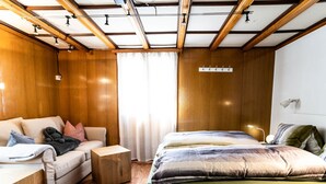 Un dormitorio moderno con techo de madera, una cama con ropa de cama verde, un sofá con varios cojines y una mesita de noche de madera.