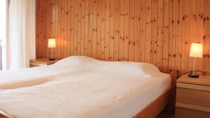 Une chambre avec un mur lambrissé en bois, un grand lit recouvert d’une couette blanche, deux tables de chevet avec des lampes et une fenêtre avec des rideaux blancs.