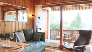Une pièce chaleureuse au décor intérieur en bois, avec un canapé, une table à manger et des chaises, ainsi qu’un balcon agrémenté de mobilier d’extérieur.