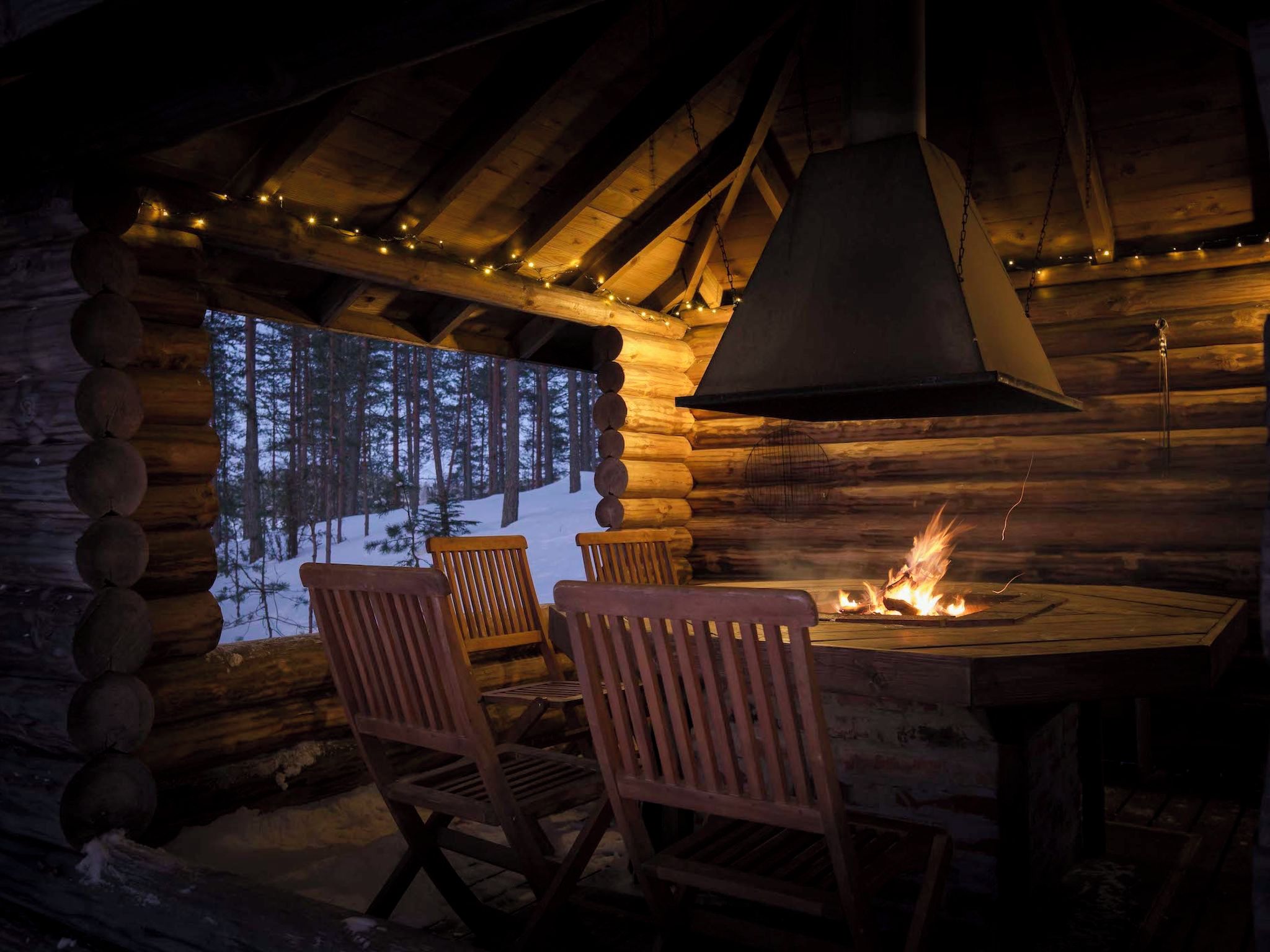 A log cabin interior with a fireplace, wooden chairs, and a view of a snowy forest through a window.
