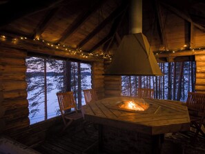 A log cabin interior with a central fire pit, wooden furniture, and a view of a forested landscape through large windows.