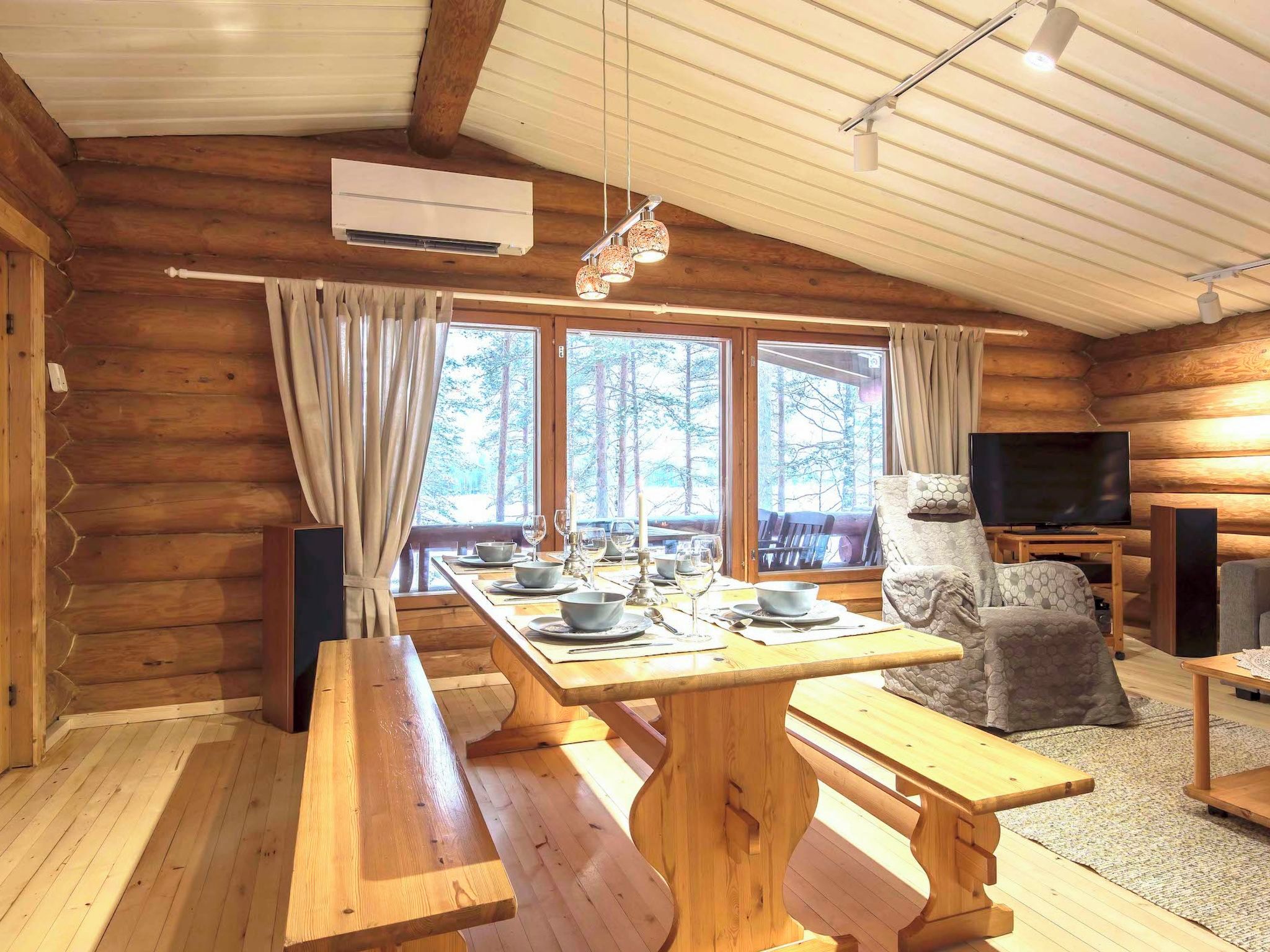 A log cabin interior with a dining table set for four, a sofa, a television, and a large window with a forest view.