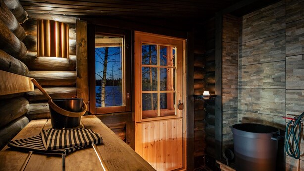 Interior acogedor de una cabaña de troncos, con una mesa de madera, una ventana que da a árboles, una puerta y un muro de piedra.