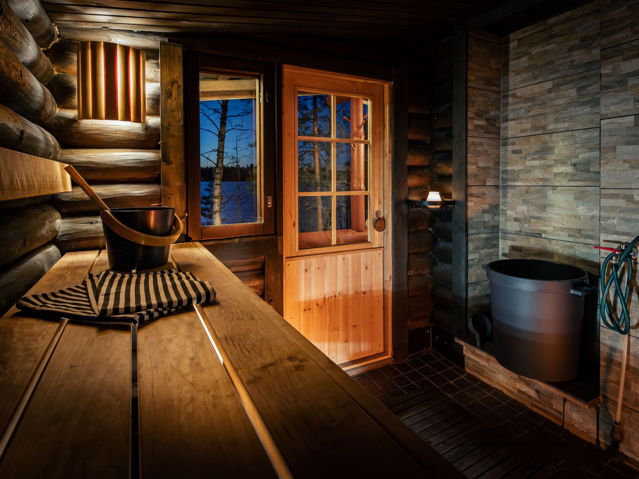 Interior de una cabaña de madera con una mesa de madera, un balde de metal, una toalla a rayas, una puerta con ventanas y una pared de piedra.