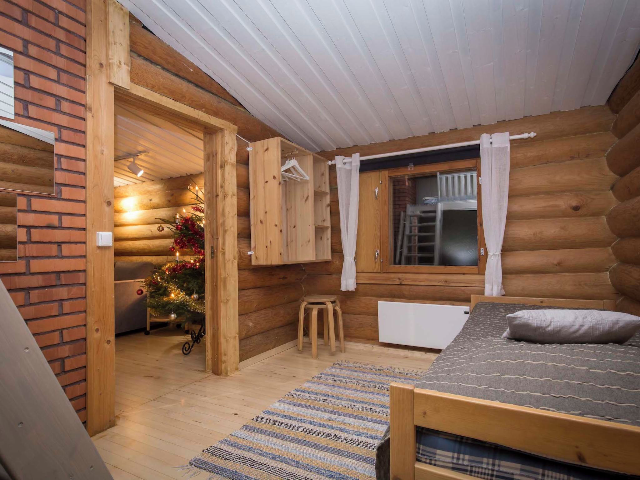 A log cabin bedroom with a bed, a radiator, a small stool, a Christmas tree, and a window with curtains.