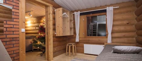 Un dormitorio de cabaña de troncos con cama, radiador, taburete pequeño, árbol de Navidad y ventana con cortinas.