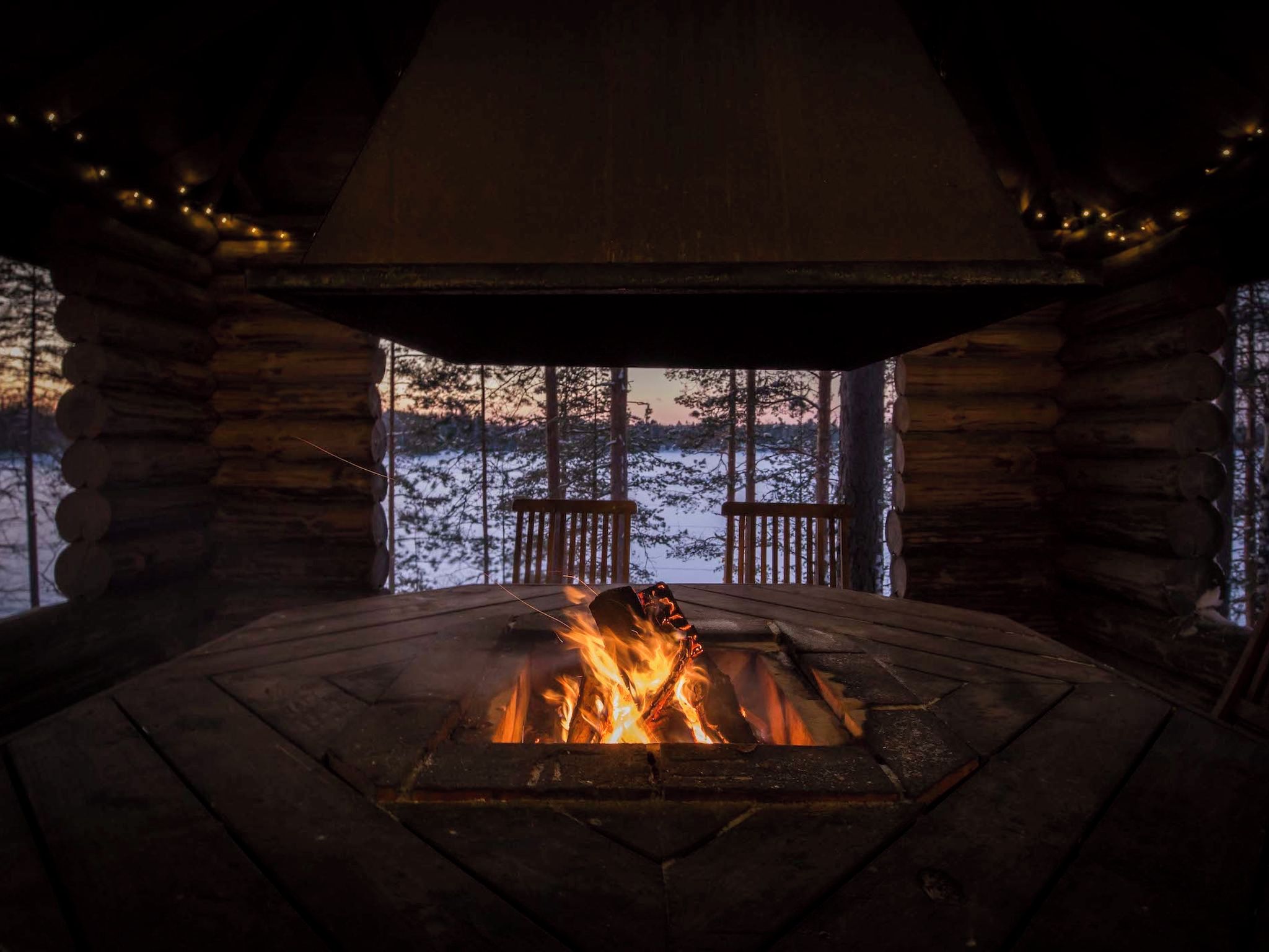 Innenansicht einer Blockhütte mit Kamin, Holzwänden und Blick auf eine verschneite Landschaft.
