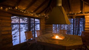 Interior acogedor de una cabaña con una chimenea central, paredes de madera y ventanas amplias con vistas a un bosque nevado.