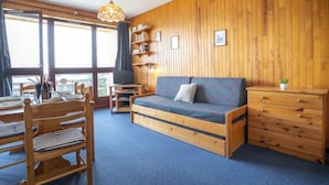 A cozy room with a wooden wall, a sofa, a TV, a bookshelf, and a dining area with chairs and a table.