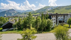 A modern hotel with a flat roof and a large parking area, surrounded by greenery and mountains in the background.
