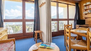 A room with large windows, a wooden bookshelf, a dining table with chairs, a small stool, and a bed.