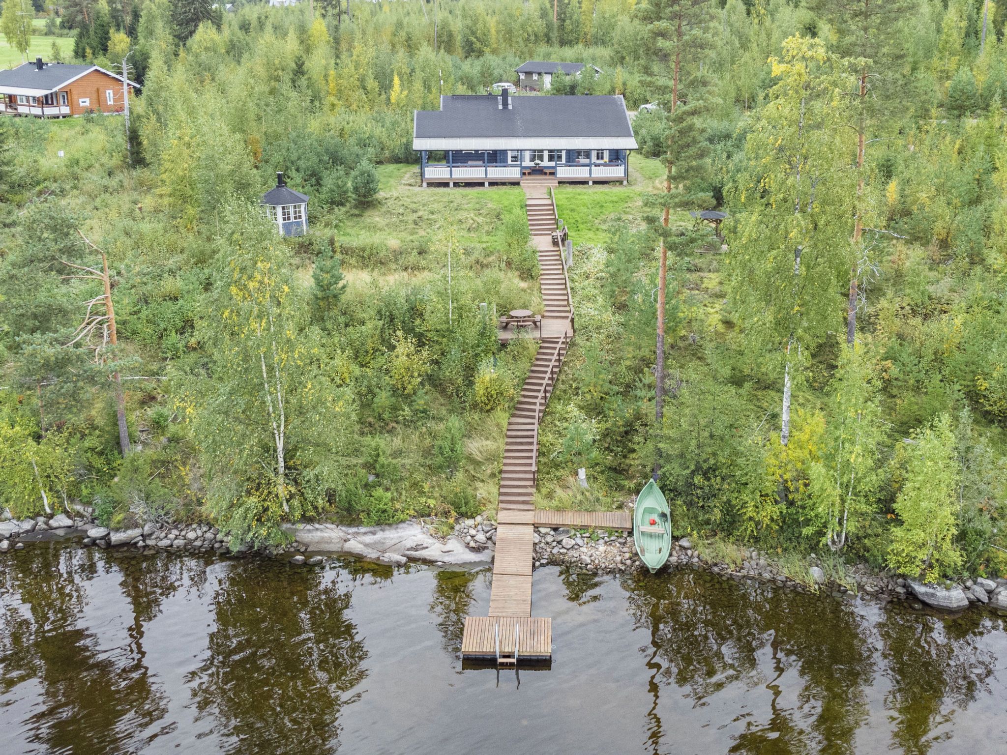 A wooden dock leading to a docked boat, surrounded by dense forest and a house in the background.