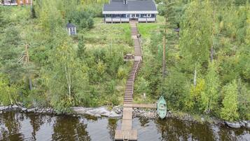 A wooden dock leading to a docked boat, surrounded by dense forest and a house in the background.