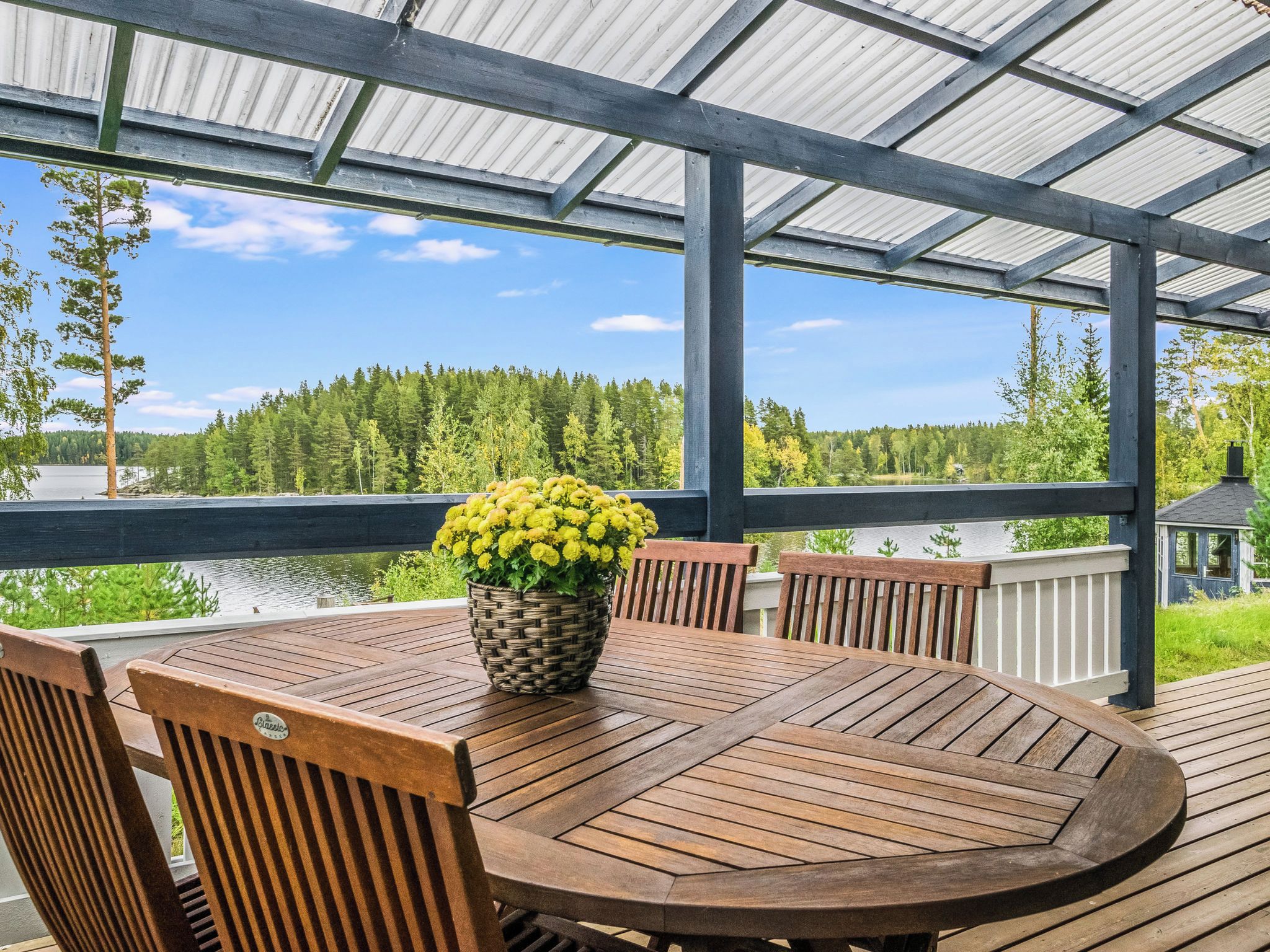 Une terrasse extérieure couverte, avec une table et des chaises en bois rondes, un pot de fleurs et une vue sur une forêt et un lac.