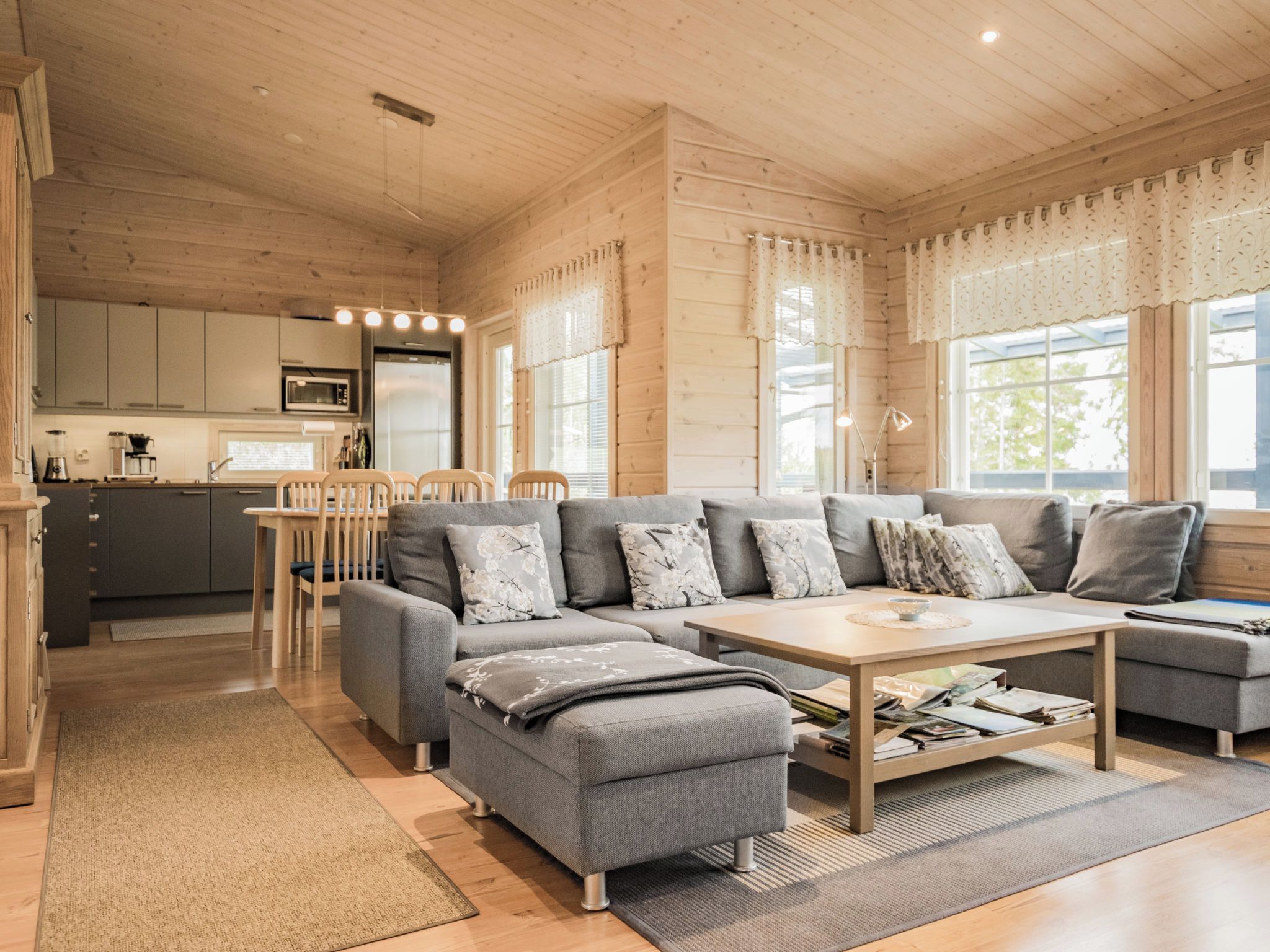 A modern log cabin interior with a grey sectional sofa, a wooden dining table, and a kitchen area with a refrigerator and microwave.