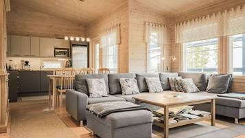A modern log cabin interior with a grey sectional sofa, a wooden dining table, and a kitchen area with a refrigerator and microwave.