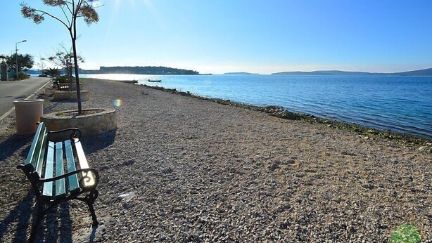 A pebble beach with a bench and a tree, overlooking a calm sea and distant hills. - Apartment Ena by Interhome (Brodarica)