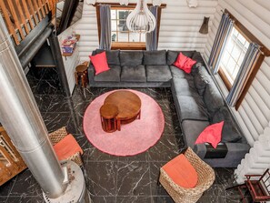 A log cabin interior with a large gray sectional sofa, a round wooden coffee table, and a pink rug.