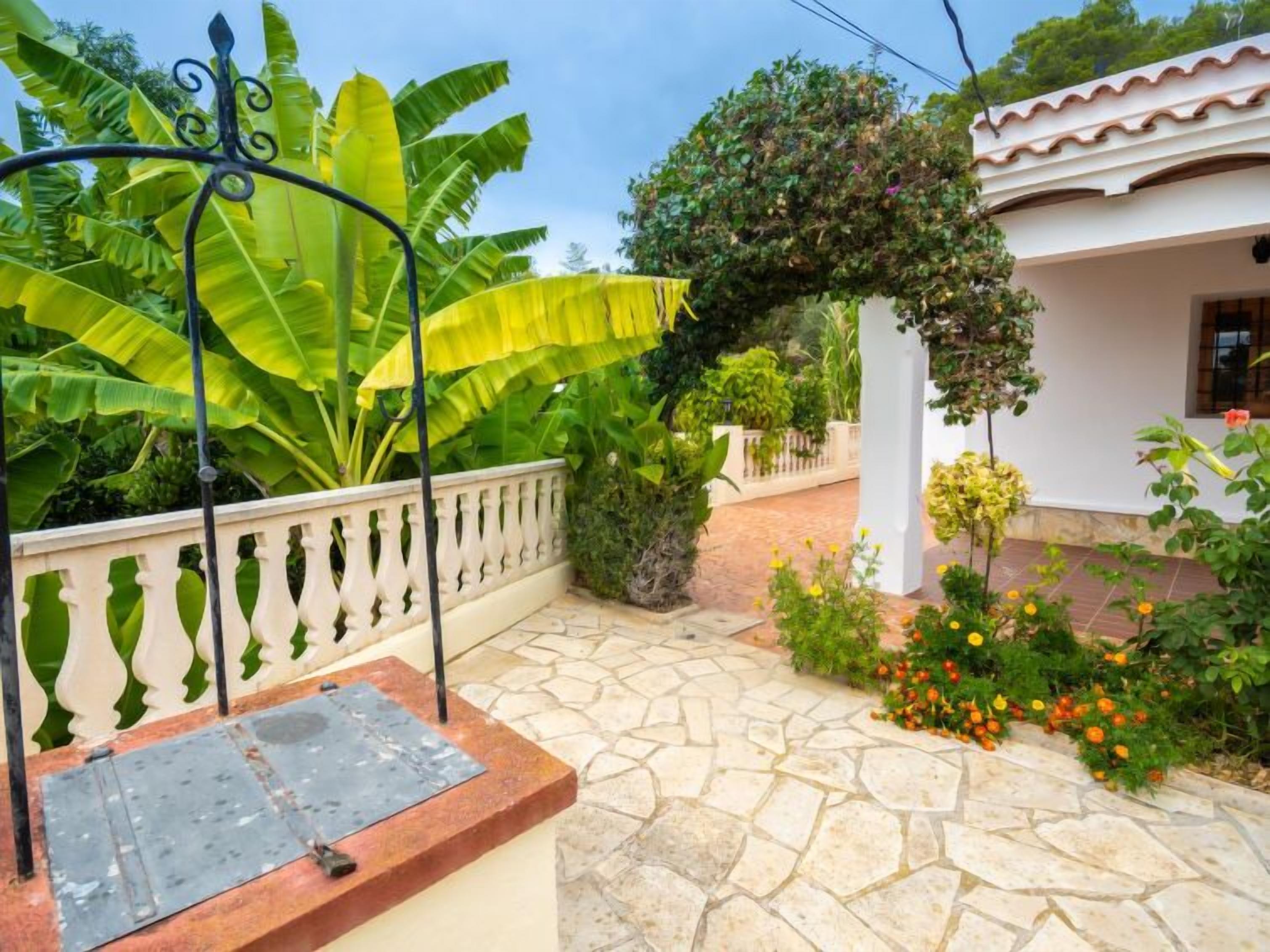 Un patio empedrado con un banco de metal, abundante vegetación y un edificio blanco con techo de tejas.