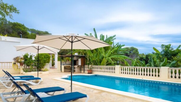 Ein Swimmingpool mit Liegestühlen und Sonnenschirmen, umgeben von einer Steindeck und Grünpflanzen.