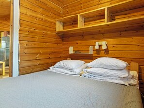 Ein Zimmer einer Holzhütte mit einem Bett, Kissen, einem Regal und einem Fenster.