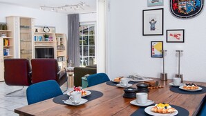 Un'area pranzo con tavolo in legno, sedie blu e piatti di cibo. Si vedono scaffali e una finestra sullo sfondo.