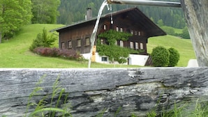 Una casa de madera rústica con techo inclinado, rodeada de abundante vegetación y un paisaje de colinas boscosas.