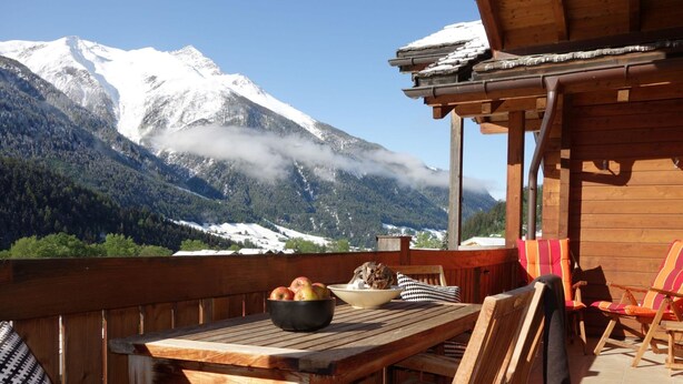 A wooden deck with a table, chairs, and a bowl of apples. A mountain with snow-covered peaks and a forested valley is visible in the background. - LuxLax 2. OG Süd by Interhome (Lax)