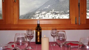 A dining table with wine glasses, a candle, and a bottle of wine, set against a backdrop of a snowy mountain view through a window. - LuxLax 2. OG Süd by Interhome (Lax)