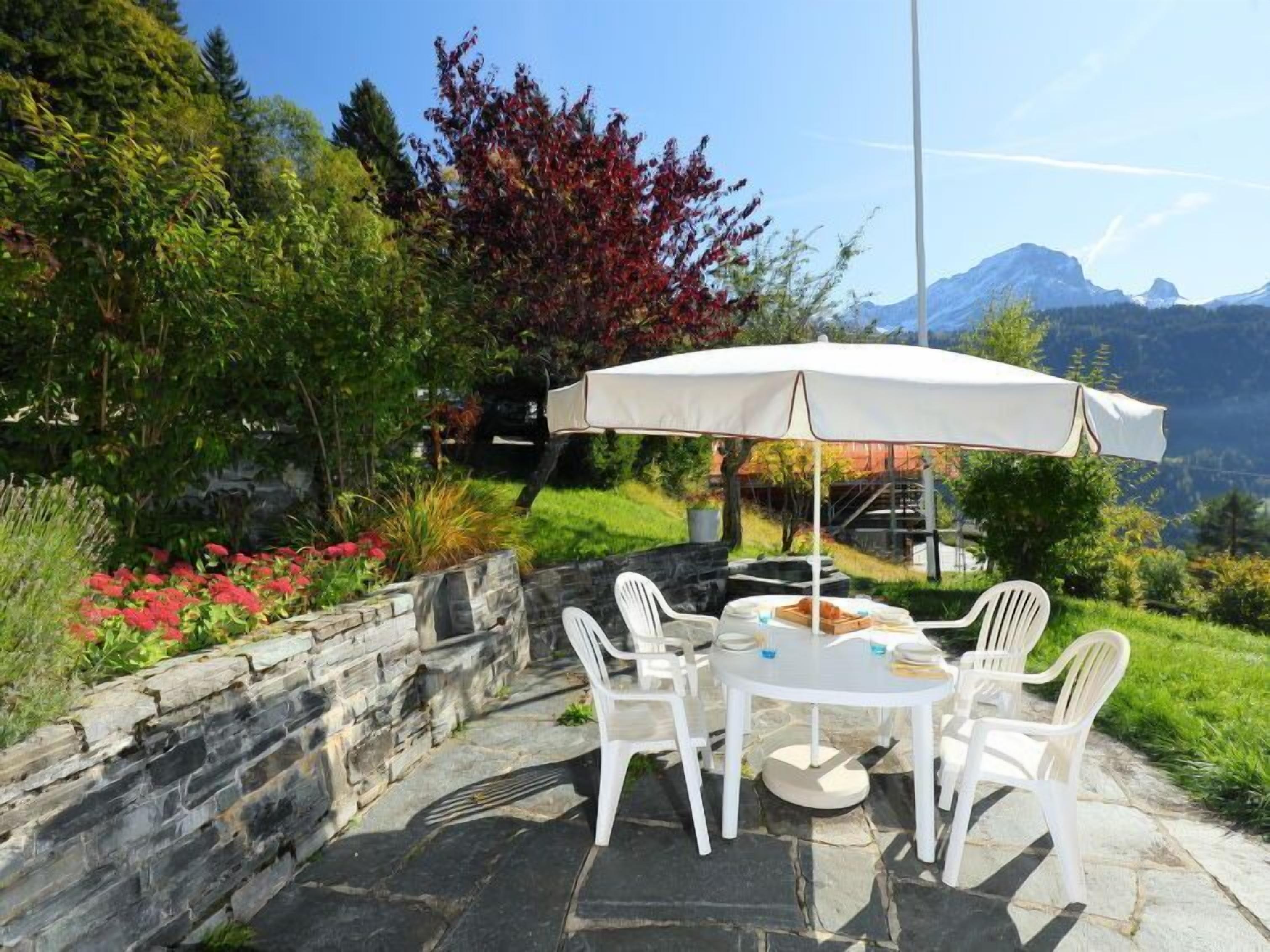 Una terrazza esterna con un muro in pietra, sedie in plastica bianca, un tavolo rotondo e un ombrellone bianco.