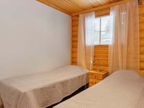 A room with two beds, wooden walls, a nightstand with a lamp, and a window with sheer curtains.