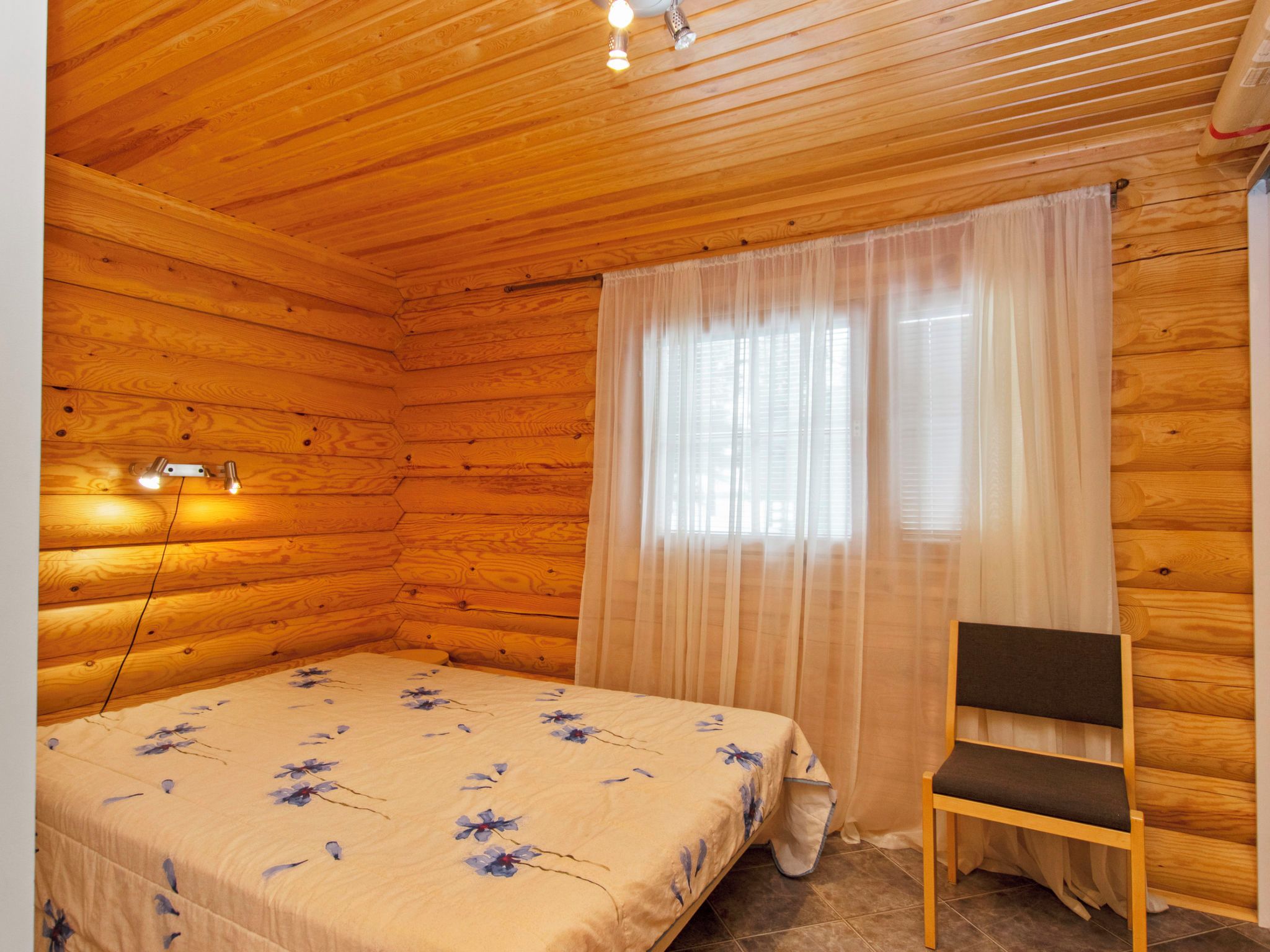 A wooden cabin bedroom with a bed, a chair, and a window with sheer curtains.