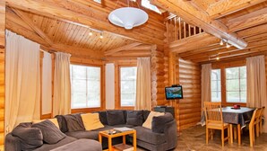 A log cabin interior with a sectional sofa, a dining table with chairs, and a television on a stand.