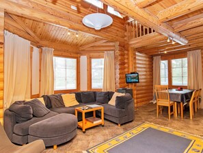 A cozy log cabin interior with a sectional sofa, a dining table with chairs, and a television on a wooden wall.