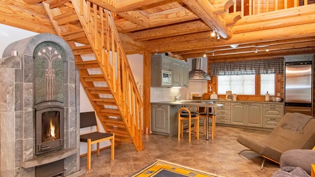 A cozy cabin interior with a stone fireplace, wooden staircase, kitchen area, and a comfortable seating area.