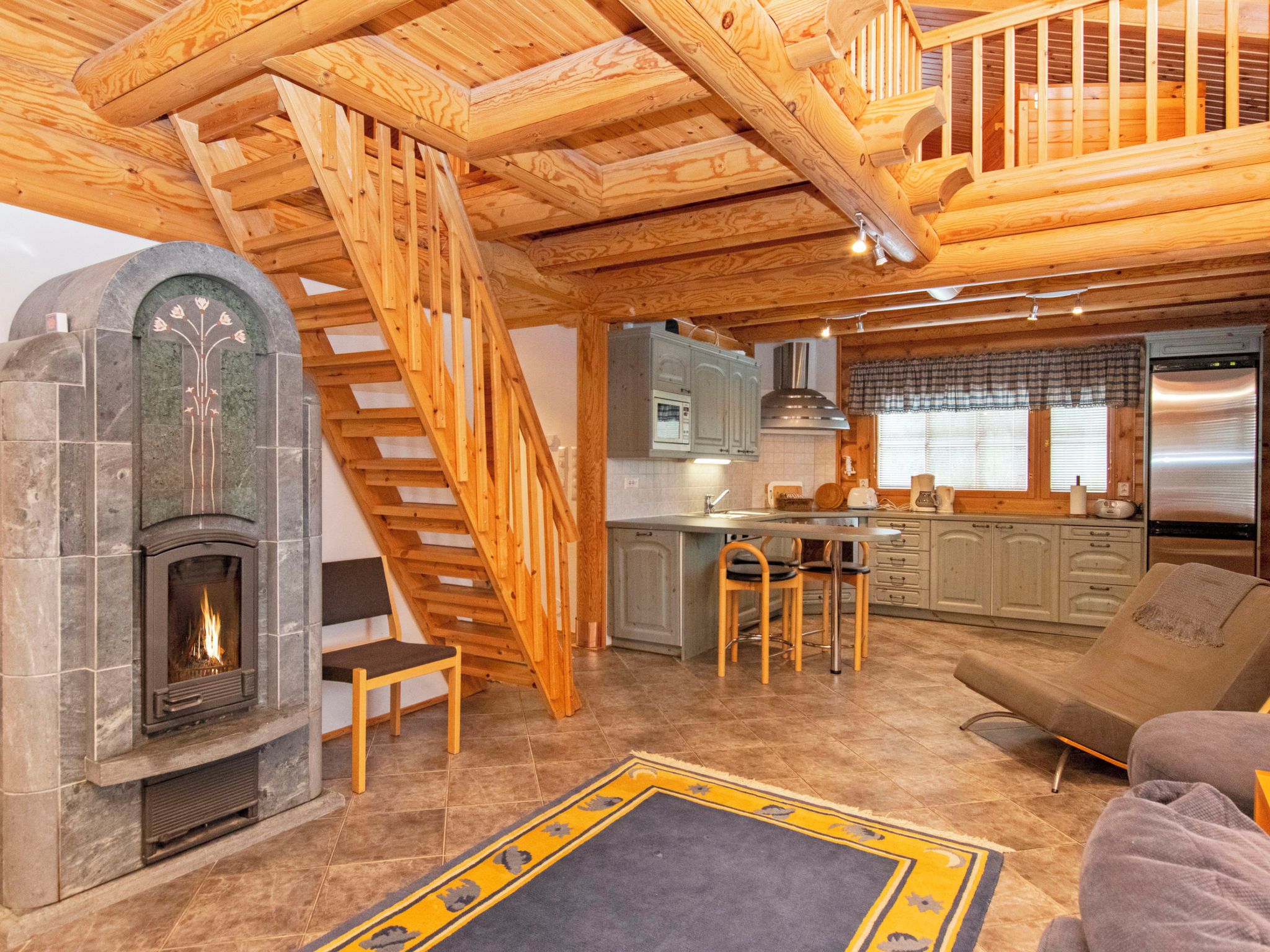 A cozy cabin interior with a fireplace, wooden staircase, kitchen area, and a sofa.