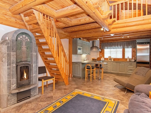 A cozy cabin interior with a fireplace, wooden staircase, kitchen area, and a sofa.
