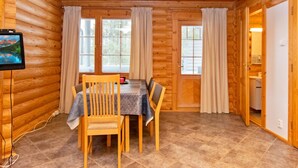 A wooden cabin interior with a dining table, chairs, curtains, and a television on the wall.