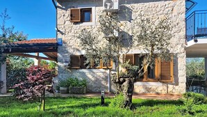 Un bâtiment en pierre avec des volets en bois, un jardin où poussent différentes espèces de plantes et un ciel bleu azur.